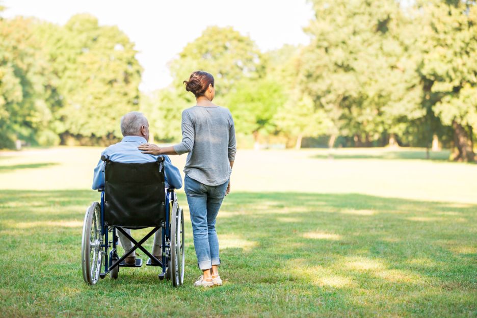 Rentnerin im Rollstuhl begleitet von einer weiblichen Pflegperson im Park, Aufnahme von hinten