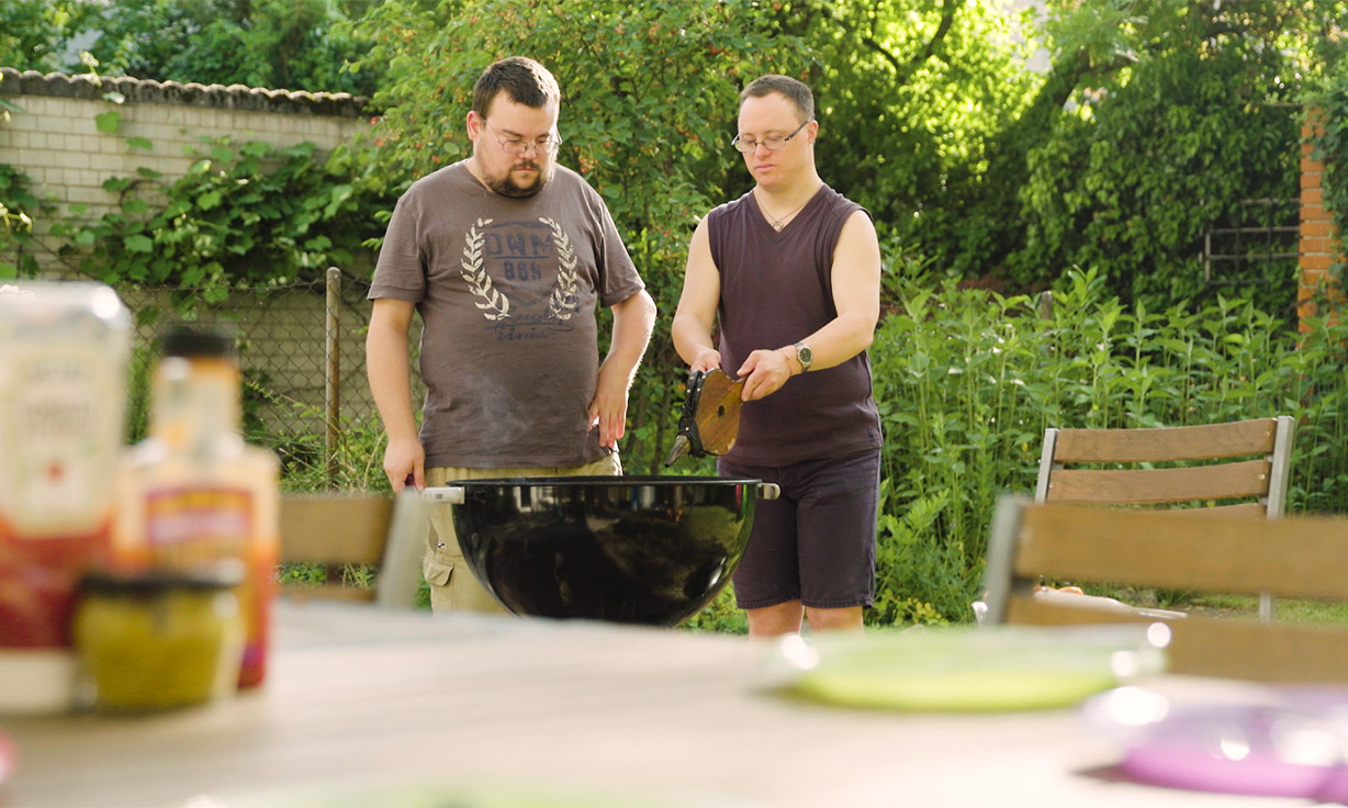 Zwei Personen grillen in einem Garten. Die eine Person bedient einen Blasbalg.