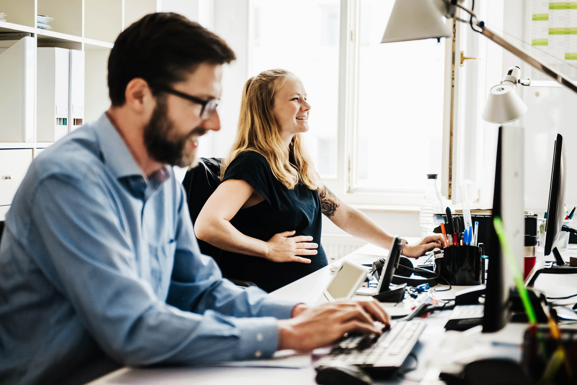 Wir gestalten ein angenehmes Arbeitsklima für unsere Mitarbeiter. Eine schwangere Frau und ihr Arbeitskollege sitzen im Büro an ihrem Arbeitsplatz