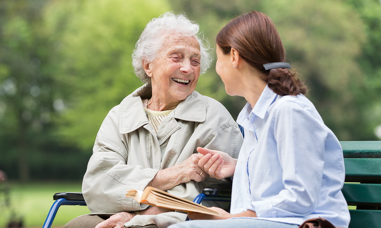 Seniorin und eine Junge Frau sitzen lachend auf einer Bank in der Natur