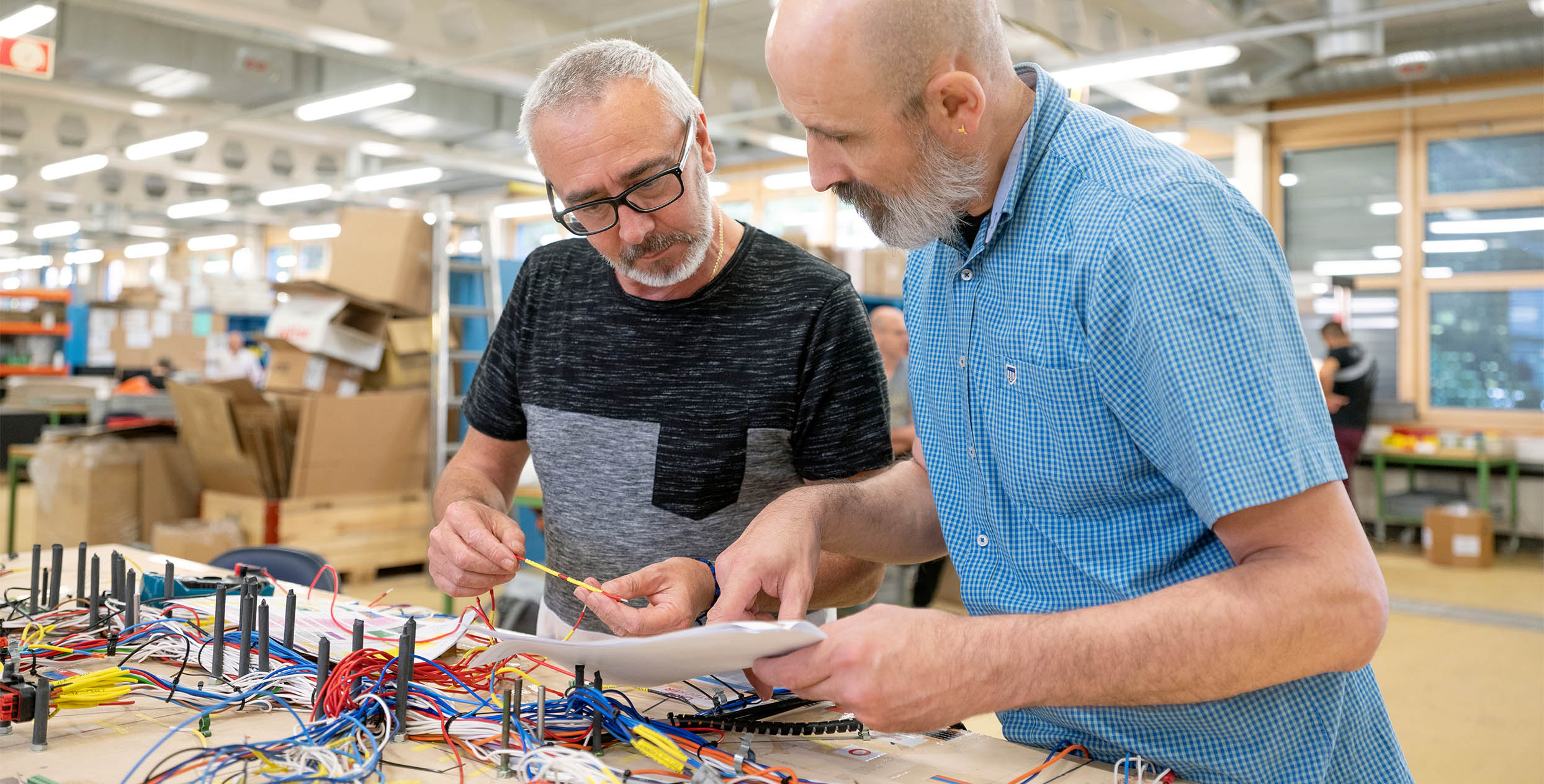 Zwei Personen stehen in einer Werkstatt neben einem Tisch, auf dem viele Kabel liegen. Die Personen unterhalten sich über ihre Arbeit