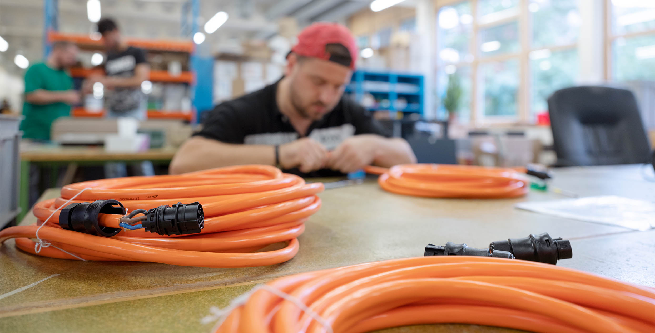 Im Vordergrund liegen aufgerollte orange Kabel auf einem Tisch, im Hintergrund sieht man unscharf eine Person, die am Tisch sitzt und arbeitet