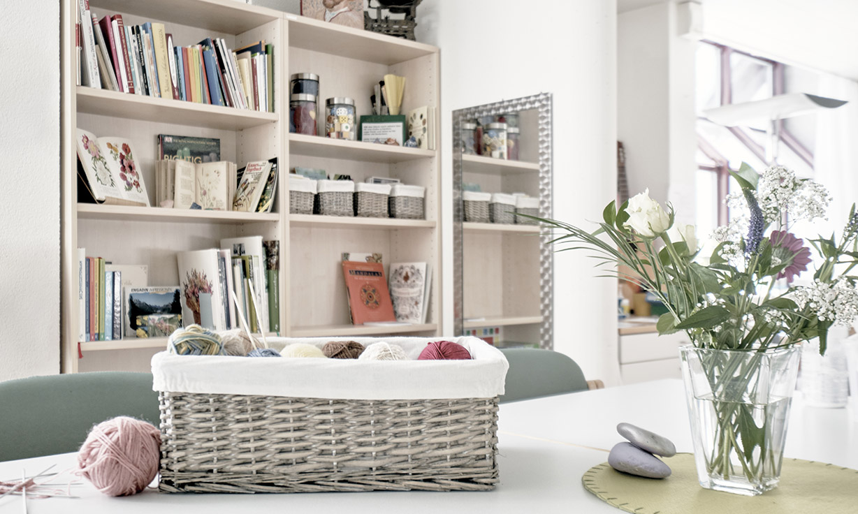 Persönlich und hell eingerichtetes Zimmer mit Strickutensilien und Blumenstraus auf dem Tisch im Vordergrund. Im Hintergrund ein Büchergestell.