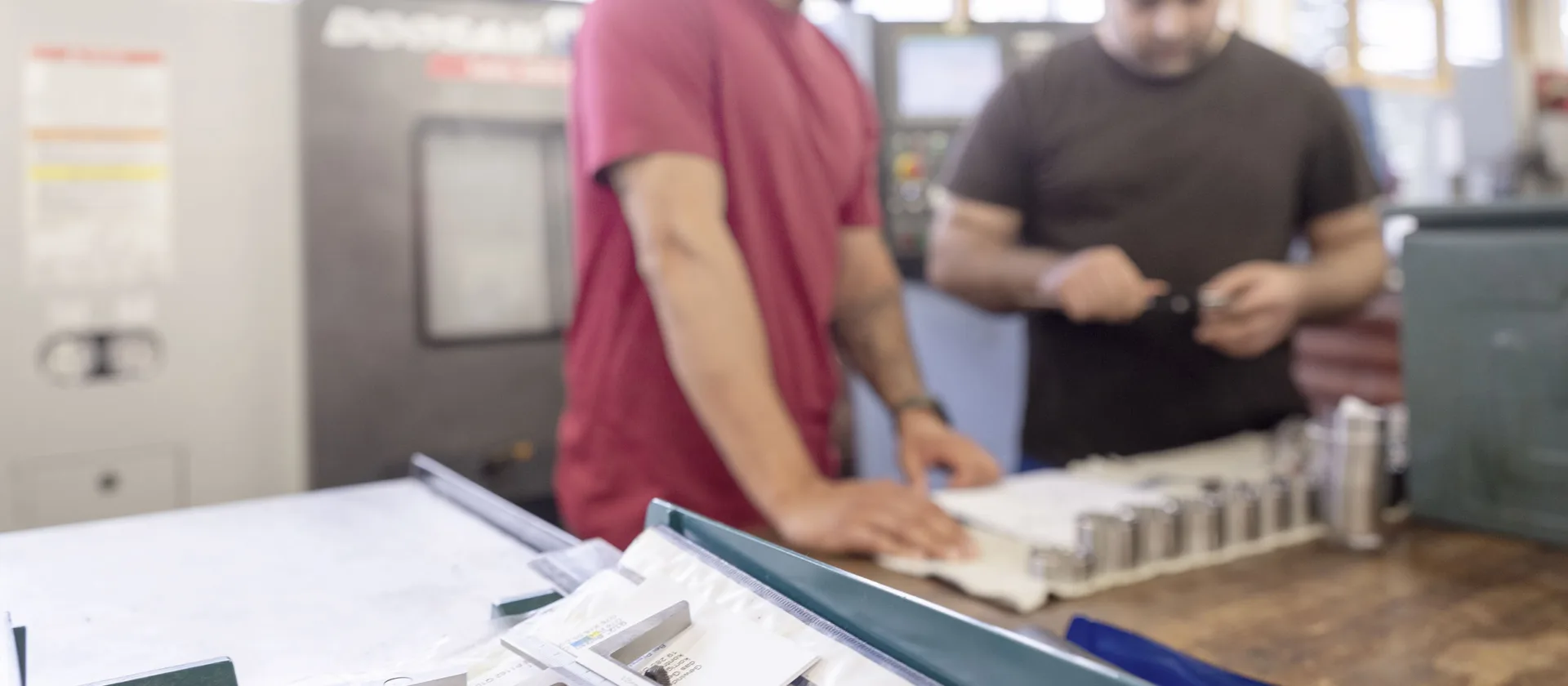 Ein Foto in einer Werkstatt. Im Hintergrund sieht man unscharf zwei Personen, die arbeiten, im Vordergrund sieht man Werkzeug liegen.