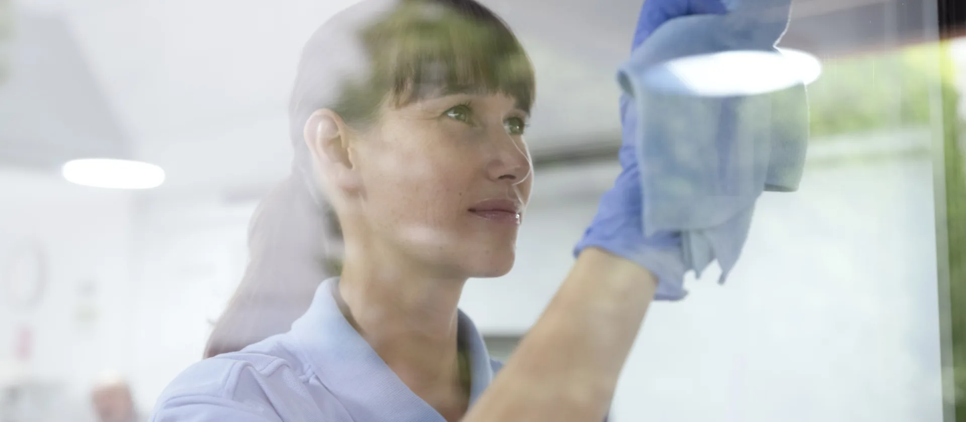 Ein Foto, das durch eine Glasscheibe aufgenommen wurde. Auf der anderen Seite vom Glas steht eine Mitarbeiterin und reinigt die Scheibe mit einem Lappen