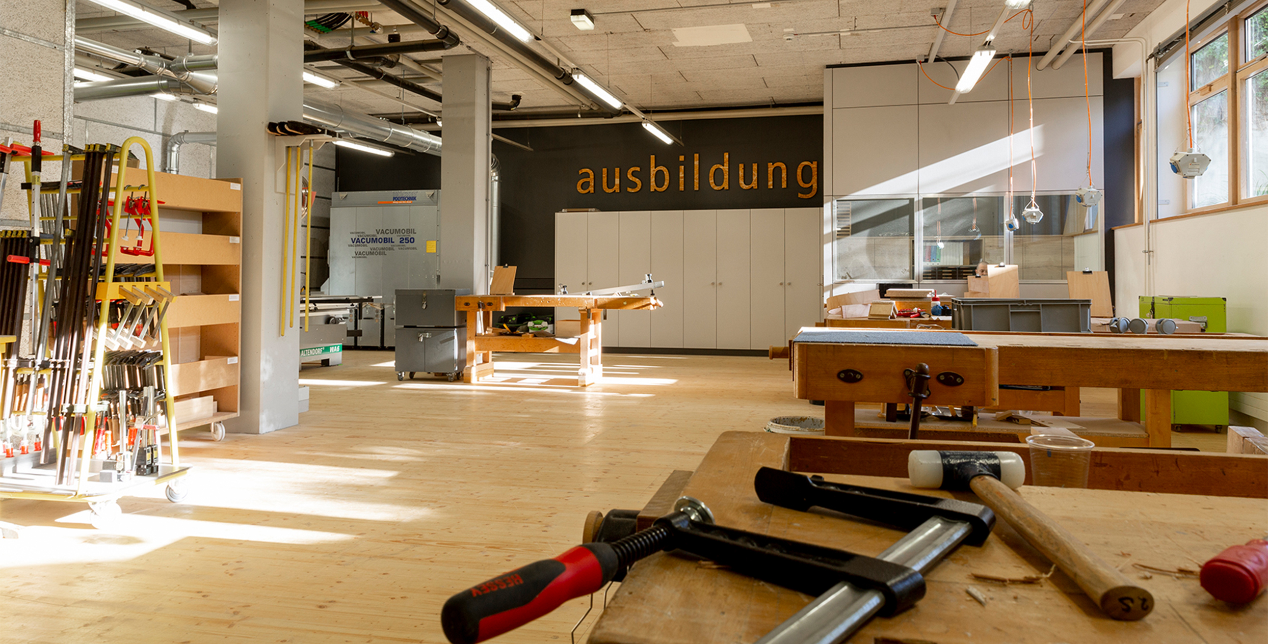 Eine Werkstatt mit mehreren Werkbänken. Im Vrdergrund sieht man eine Schraubzwinge und einen Hammer auf einer Werkbank liegen.