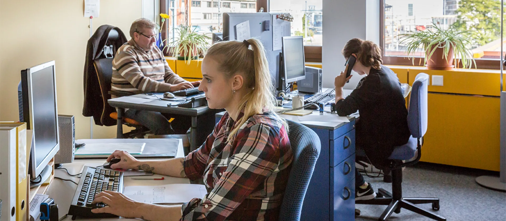 Ein Büro mit drei Schreibtischen, an denen Personen arbeiten.