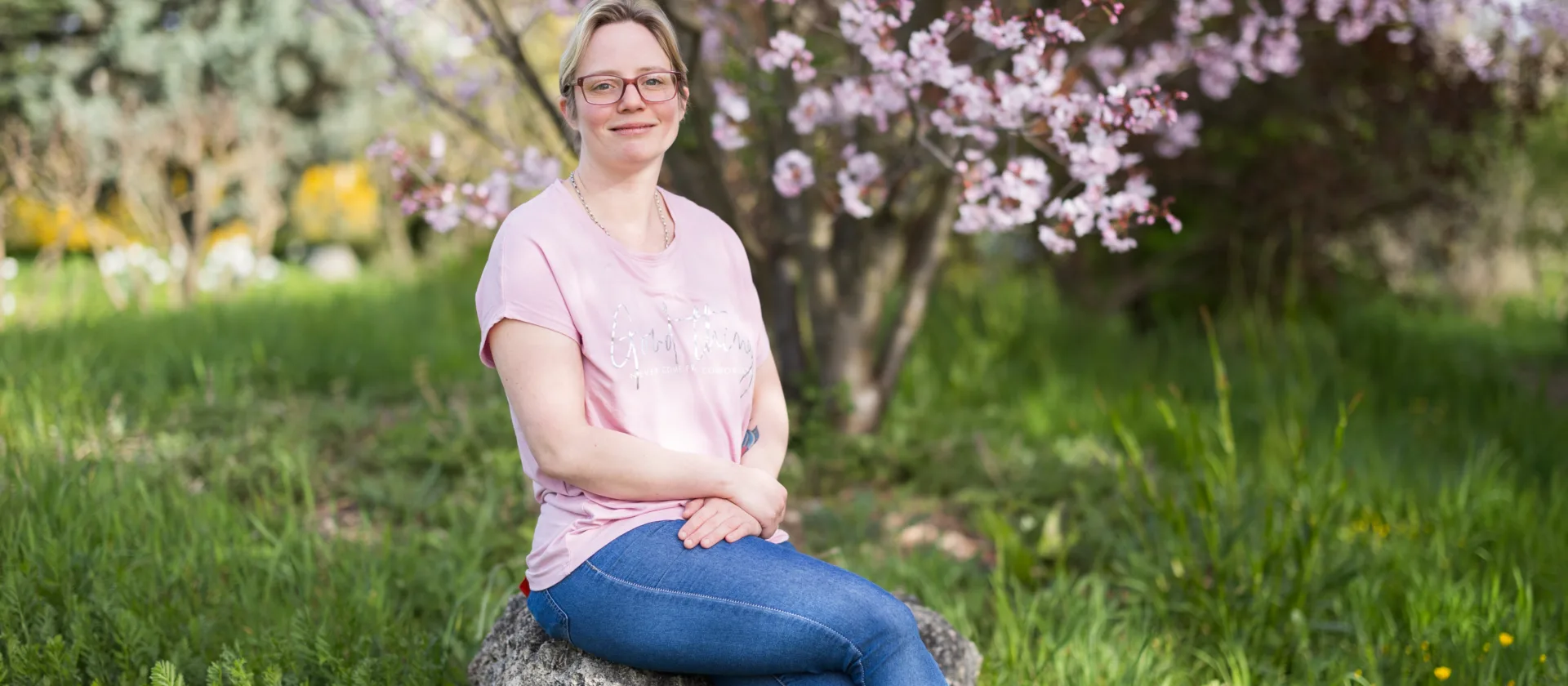 Bild einer jungen Frau auf einem Stein sitzend vor einem Garten.