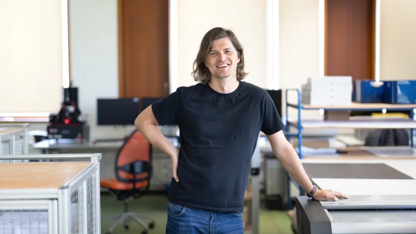 Mann in schwarzem T-Shirt steht lächelnd im Büro neben einem Gerät.