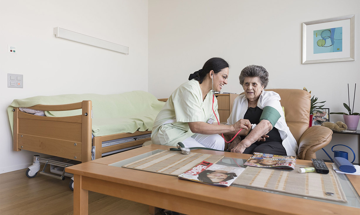 Fachfrau Betreuung untersucht eine Person, die auf einem Sofa sitzt.