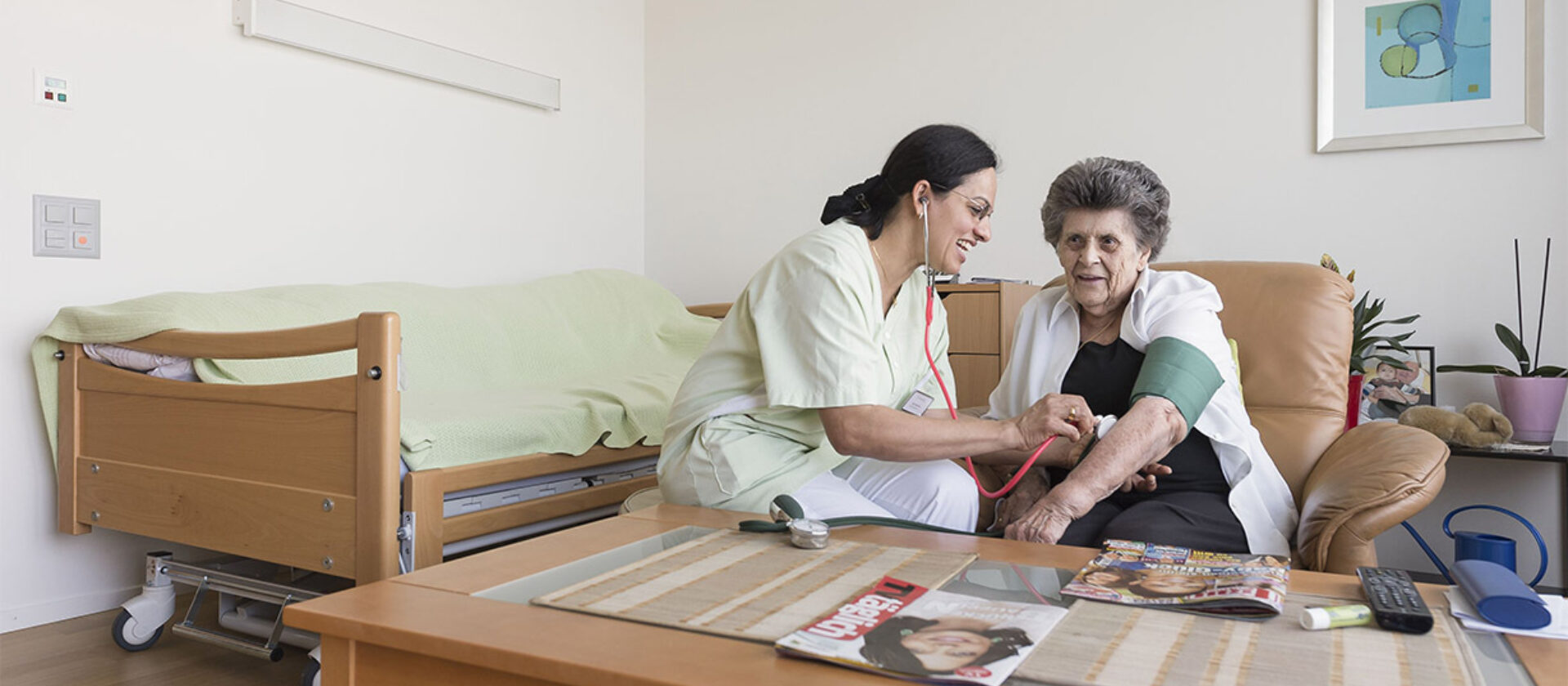 Fachfrau Betreuung untersucht eine Person, die auf einem Sofa sitzt.