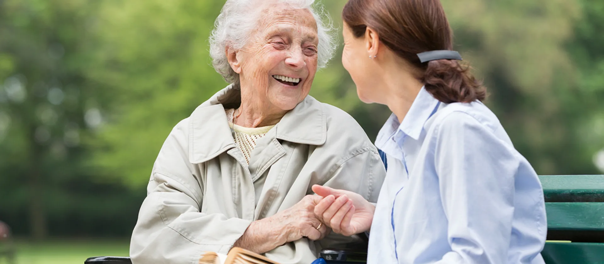 Seniorin und eine Junge Frau sitzen lachend auf einer Bank in der Natur