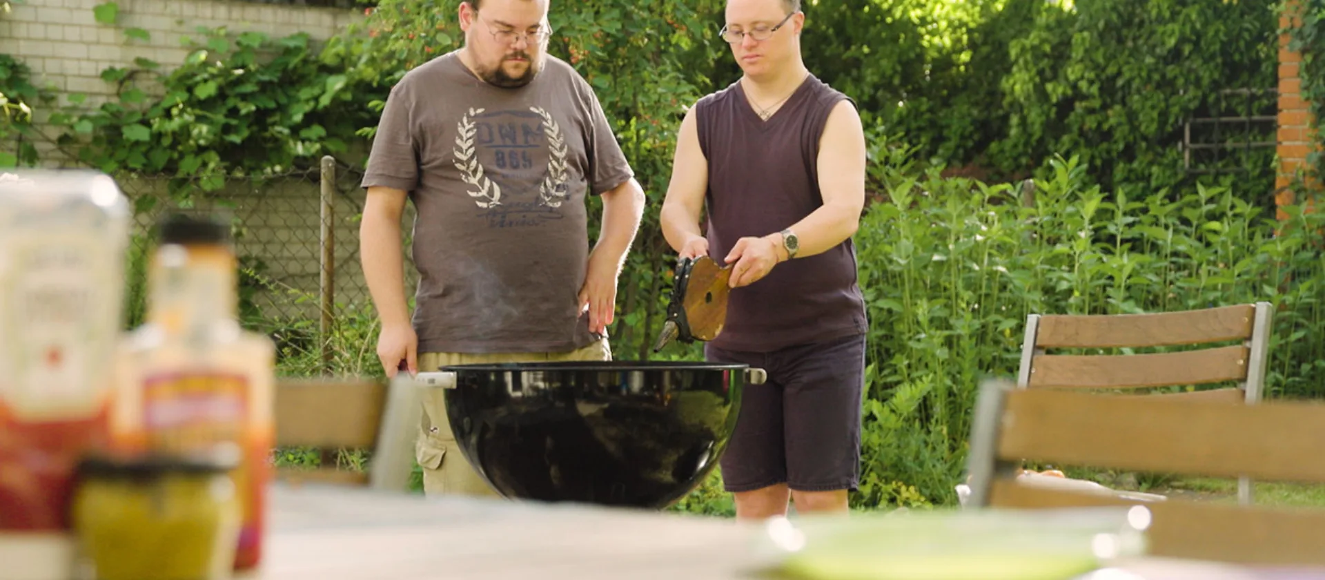 Zwei Personen grillen in einem Garten. Die eine Person bedient einen Blasbalg.