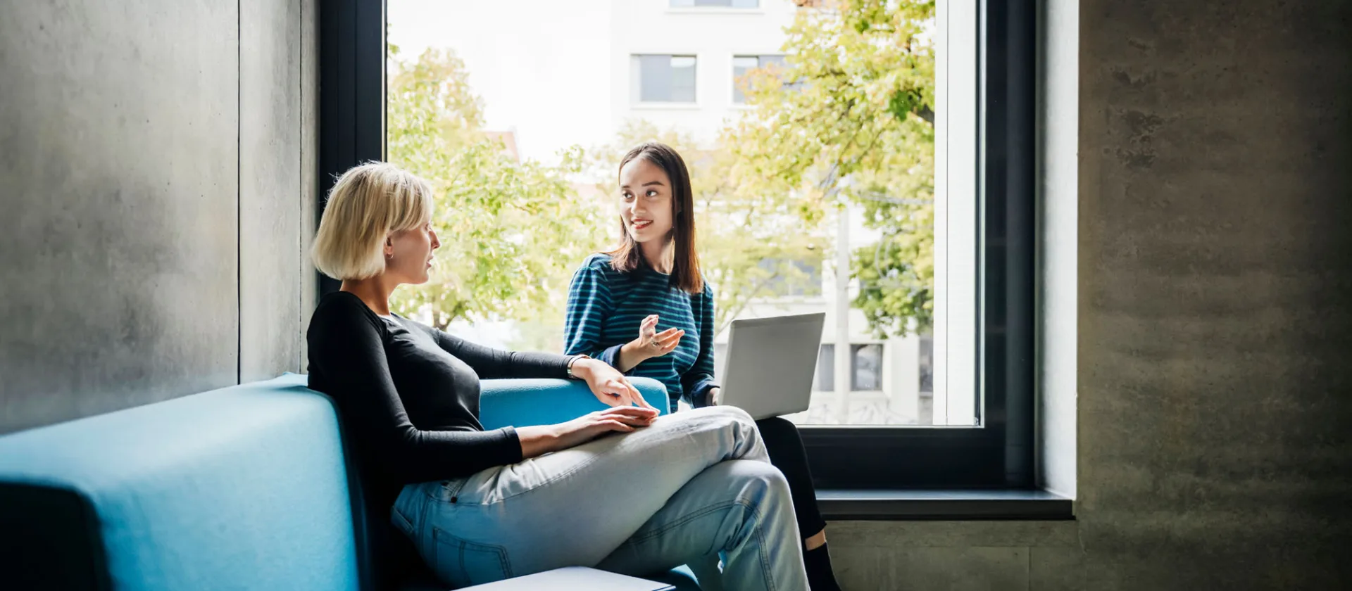 Eine Lernende und eine Ausbildnerin im Gespräch auf dem Sofa vor einem grossem Fenster.