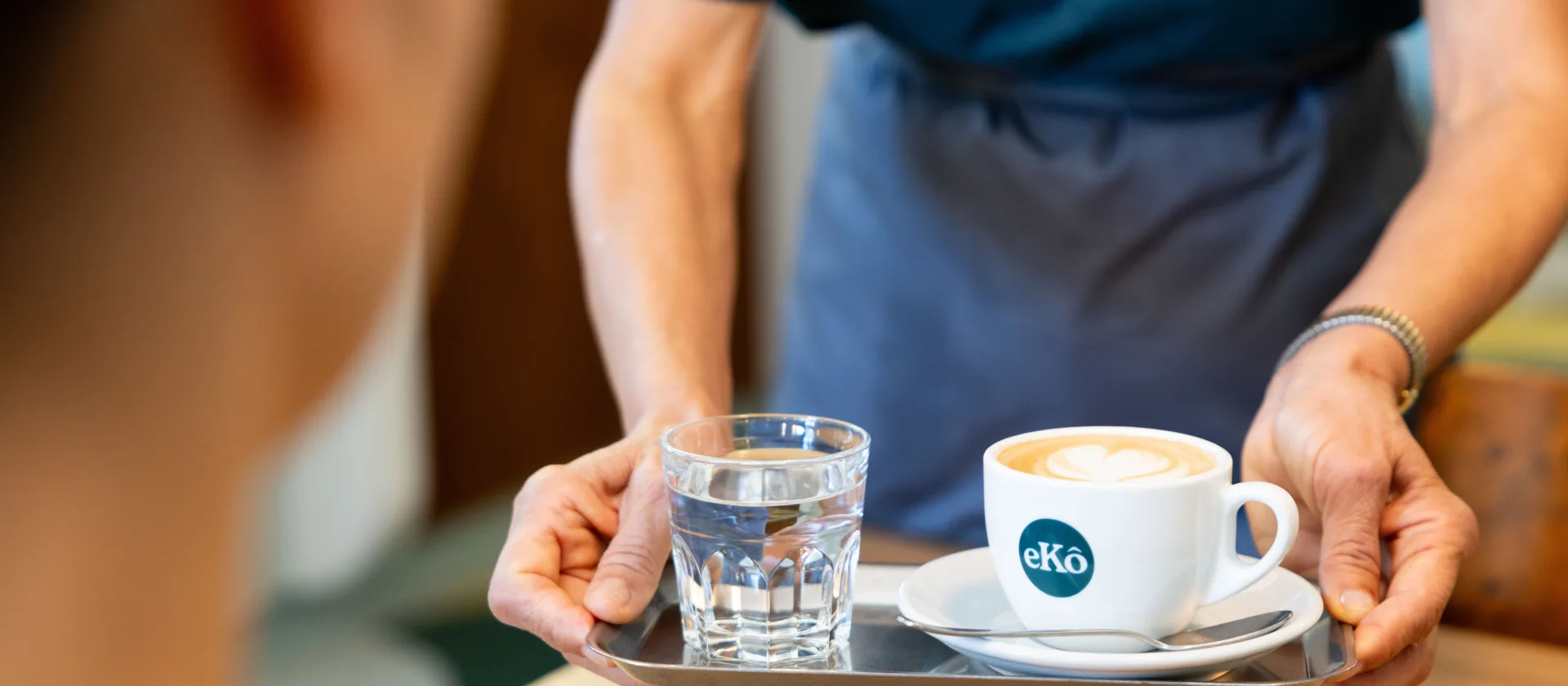 Ein Tablett mit einem Cappuccino und einem Glas Wasser darauf. Zwei Hände halten das kleine Tablett und stellen es gleich auf den Tisch.